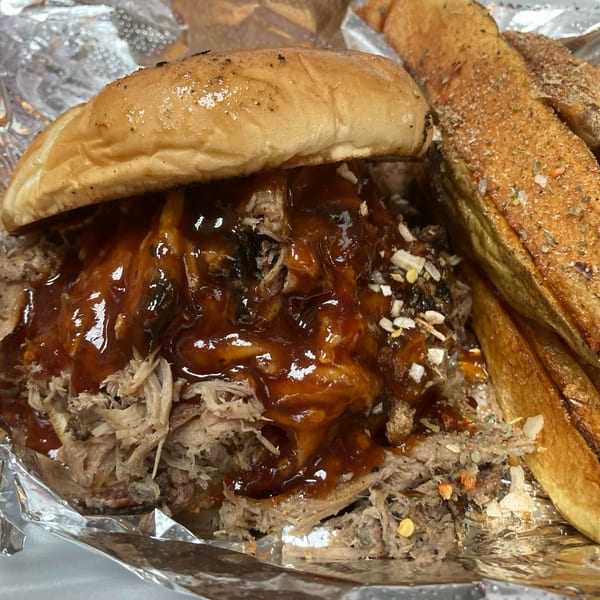 Close-up of a Five Ace's Smokestaq Lightning pulled pork sandwich with BBQ sauce and seasoned fries.
