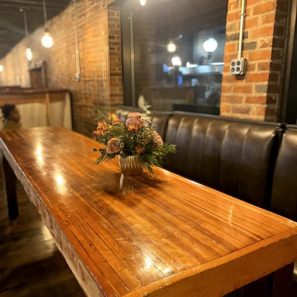 A long wooden table with a floral centerpiece, next to a dark leather booth in a dimly lit restaurant.